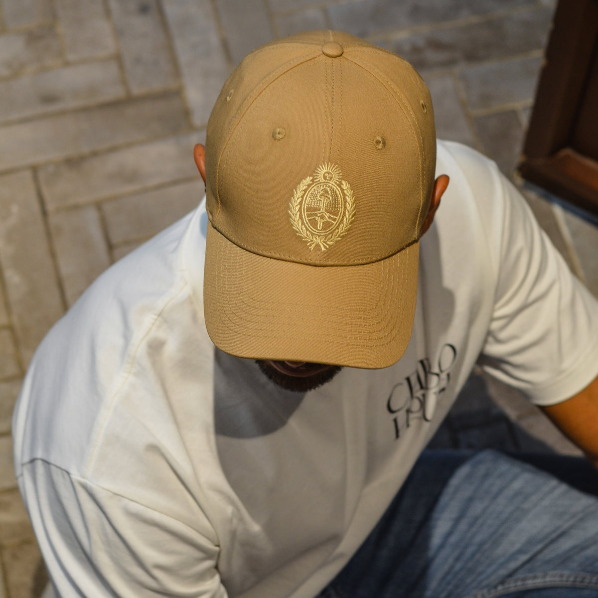 A man wearing a camel baseball cap with a gold embroidered Argentina emblem and a white t-shirt, looking down while sitting on a stone floor.