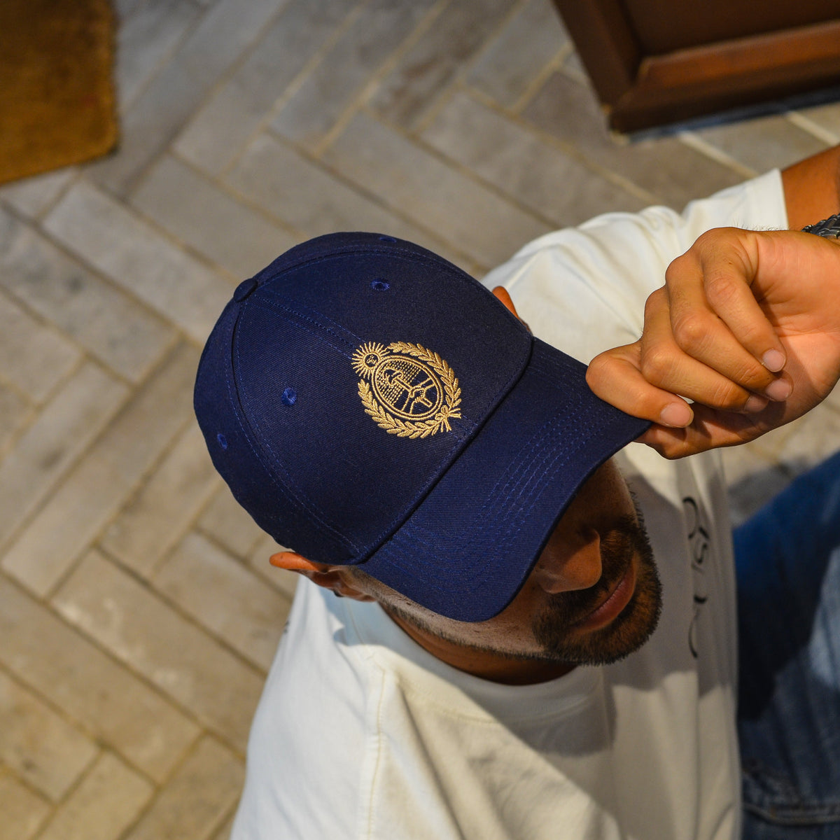 A man wearing a navy blue baseball cap with a gold embroidered argentina national emblem holding the brim of the cap with his right hand.