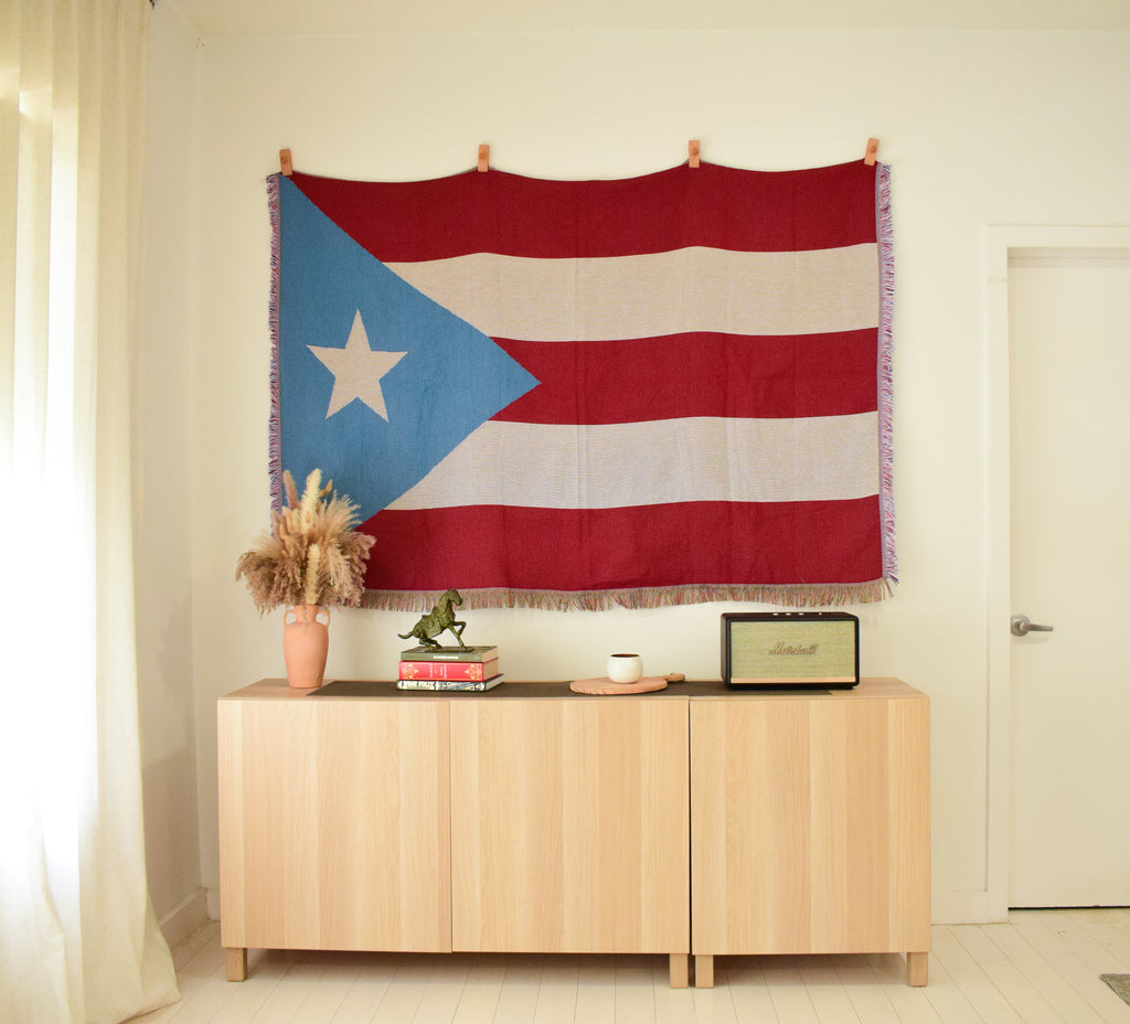 Puerto Rican flag woven throw blanket Ceibo House hangs on a cream wall above a light wood credenza in a styled room setting.