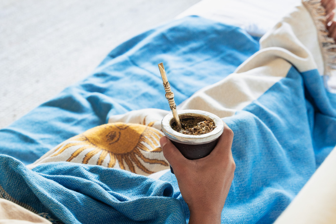 Person holding a cup of mate with an argentinian blanket and pillow in the background